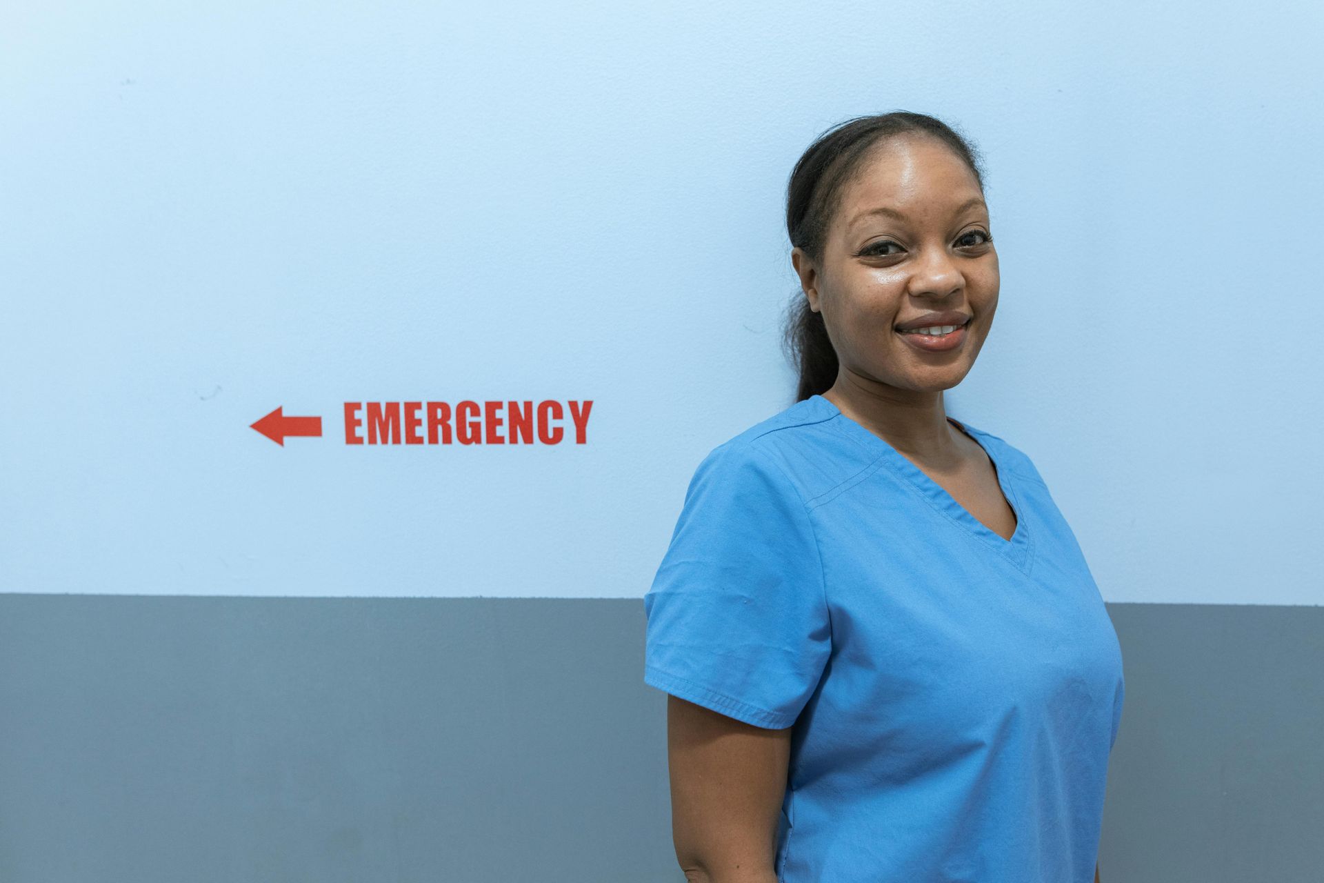 Nurse in blue scrubs smiles near an emergency exit sign.