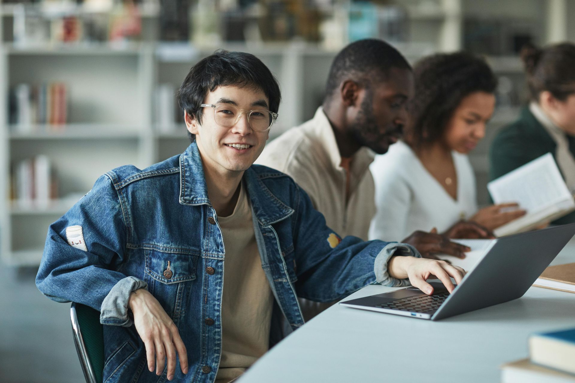 Man in a denim jacket smiles, using a laptop at a library table, with other people reading in the background.