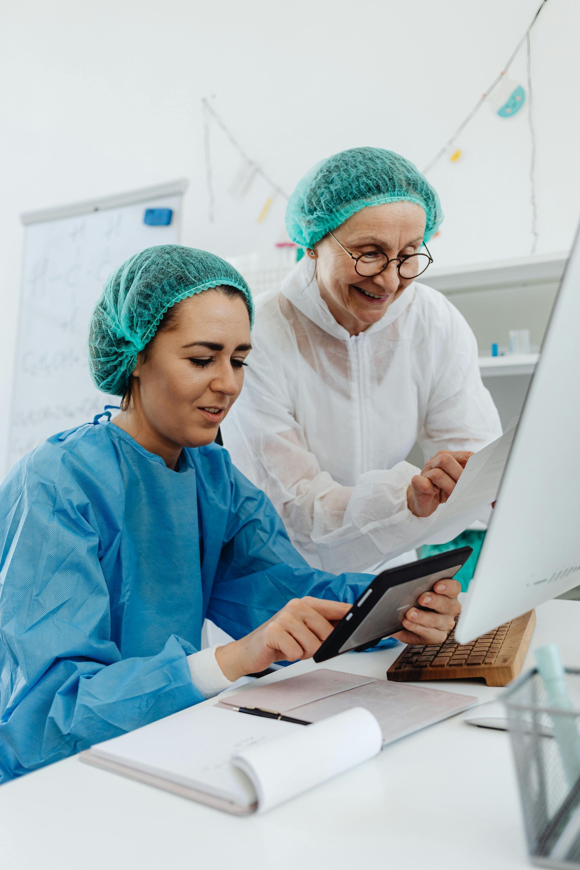 Two people in medical attire look at a tablet and paper near a computer in an office.