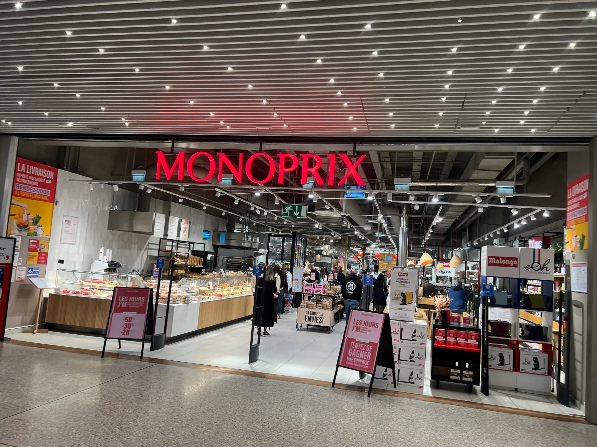 Entrée d'un supermarché Monoprix avec enseigne lumineuse rouge, comptoir de charcuterie et affiches publicitaires sur chevalets.