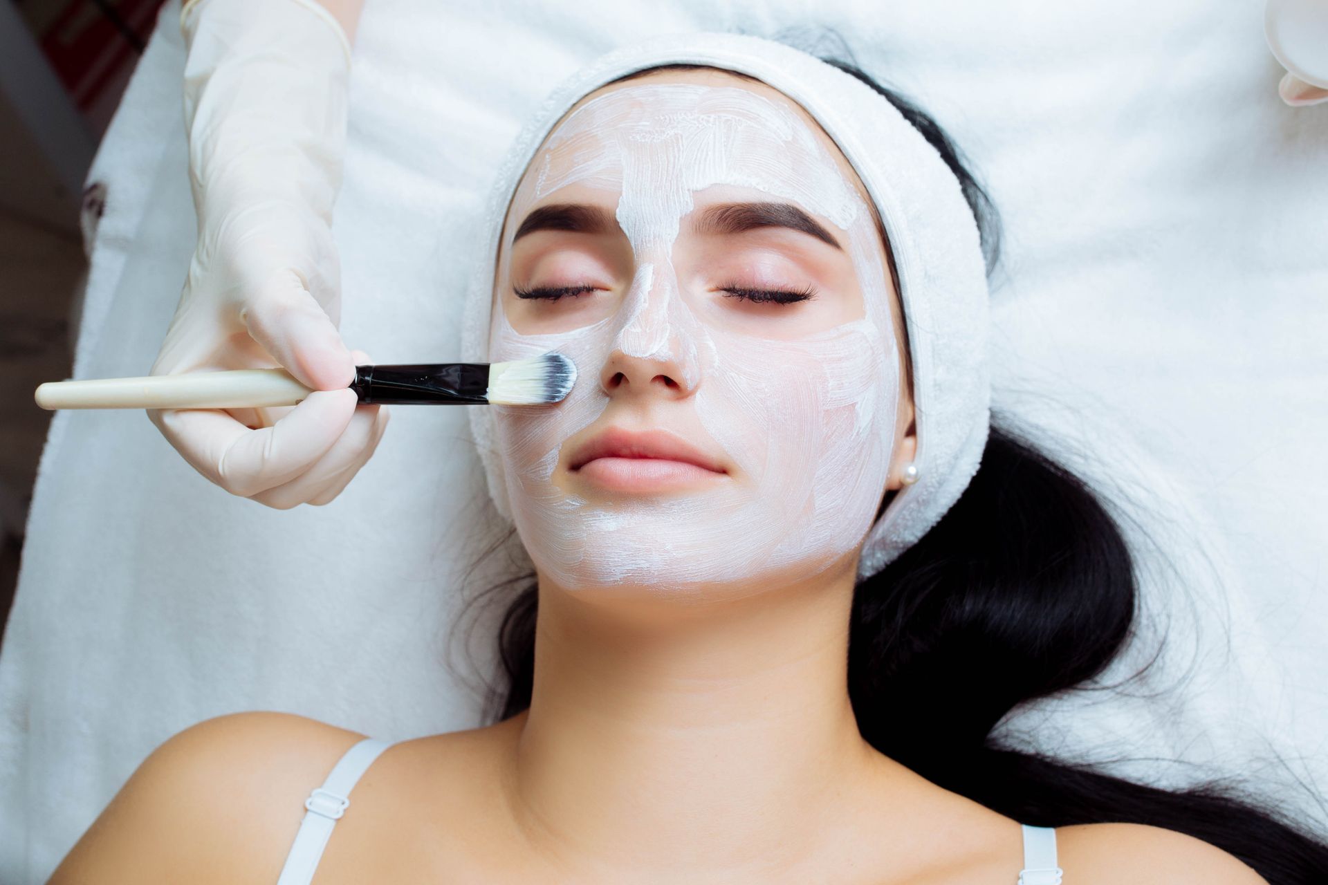 A person with a white face mask reclines while a gloved hand applies the product with a brush.