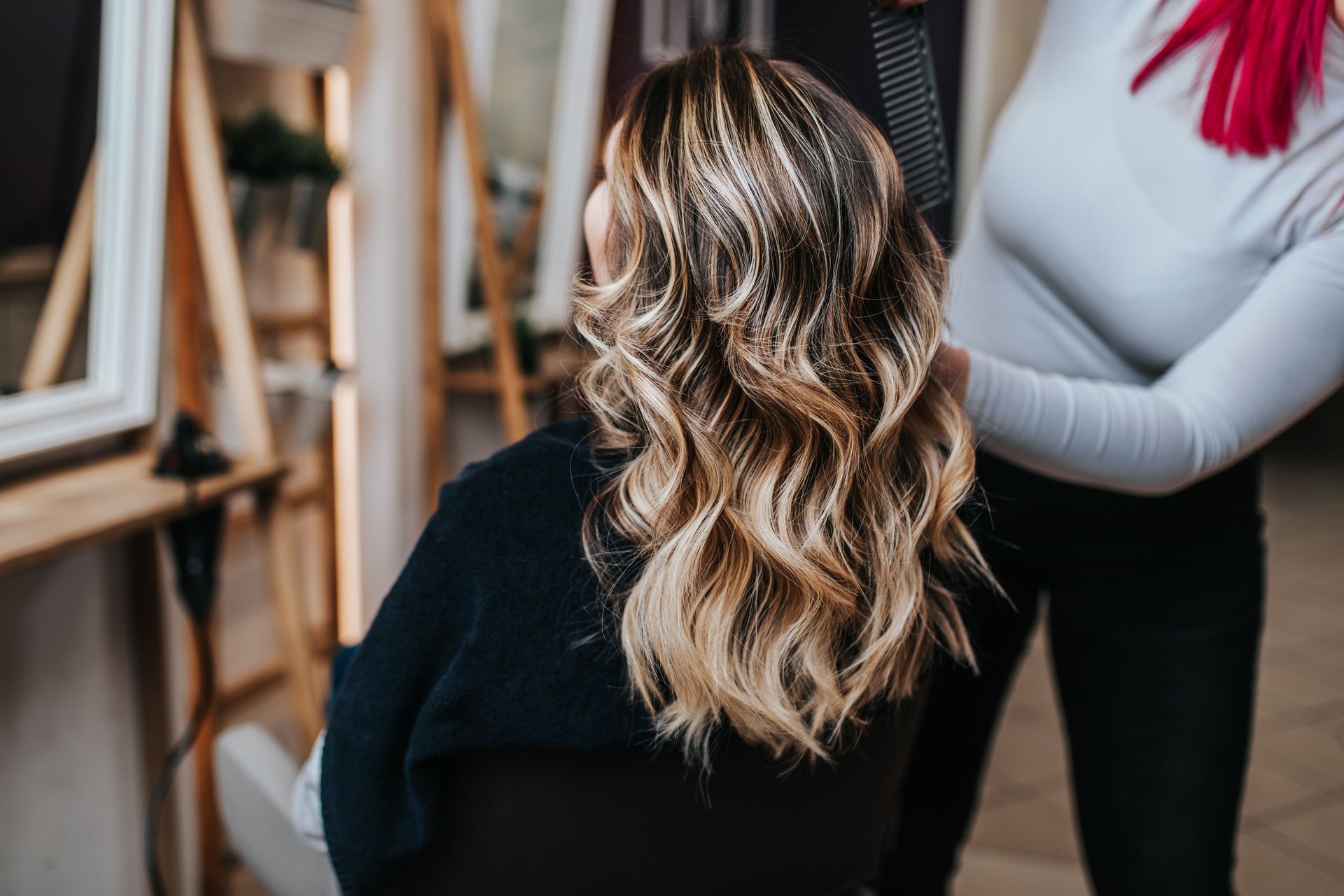 Person with wavy blonde hair seated in a salon chair while a stylist stands beside them.
