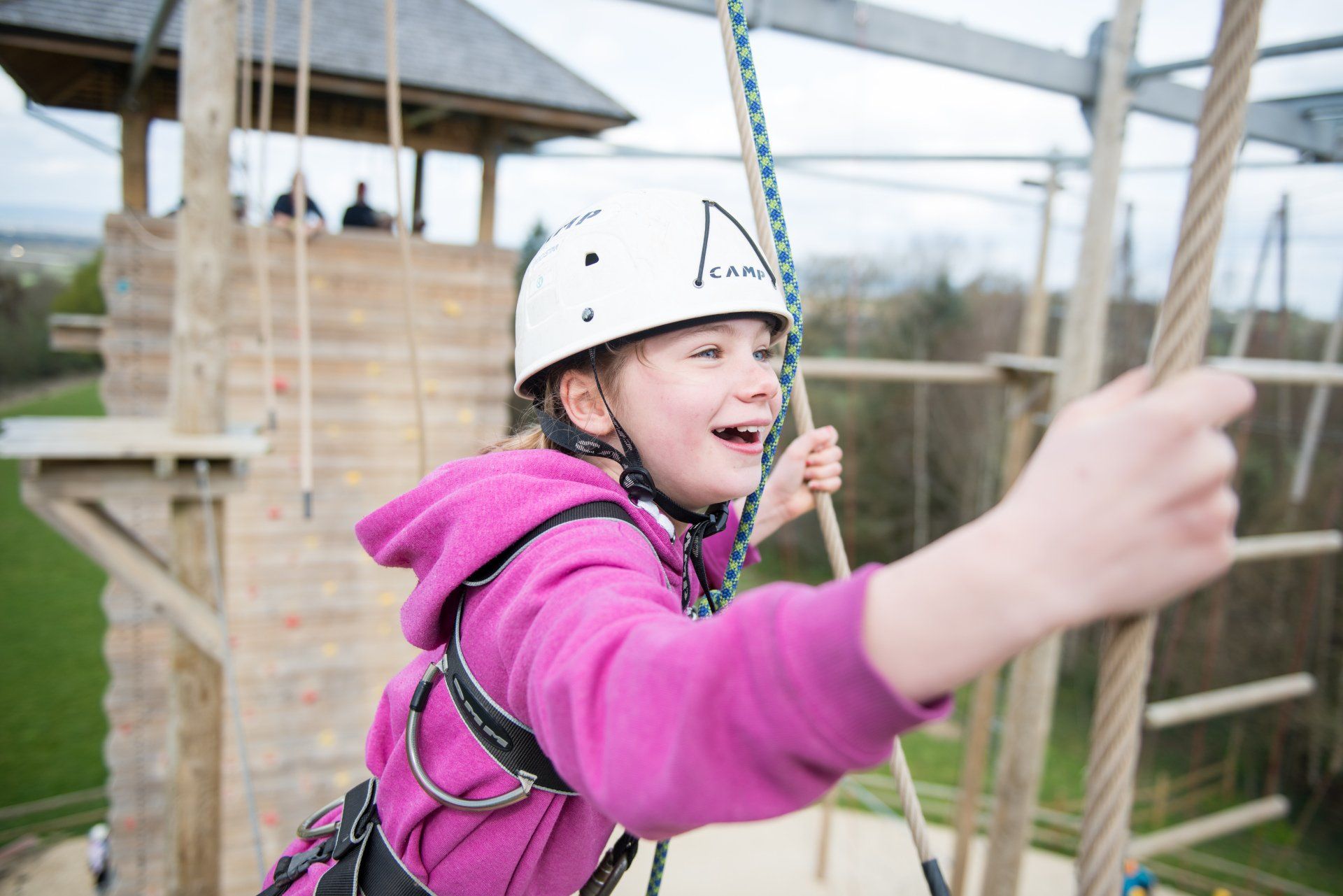 A young girl wearing a pink jacket and a white helmet is climbing a ropes course.