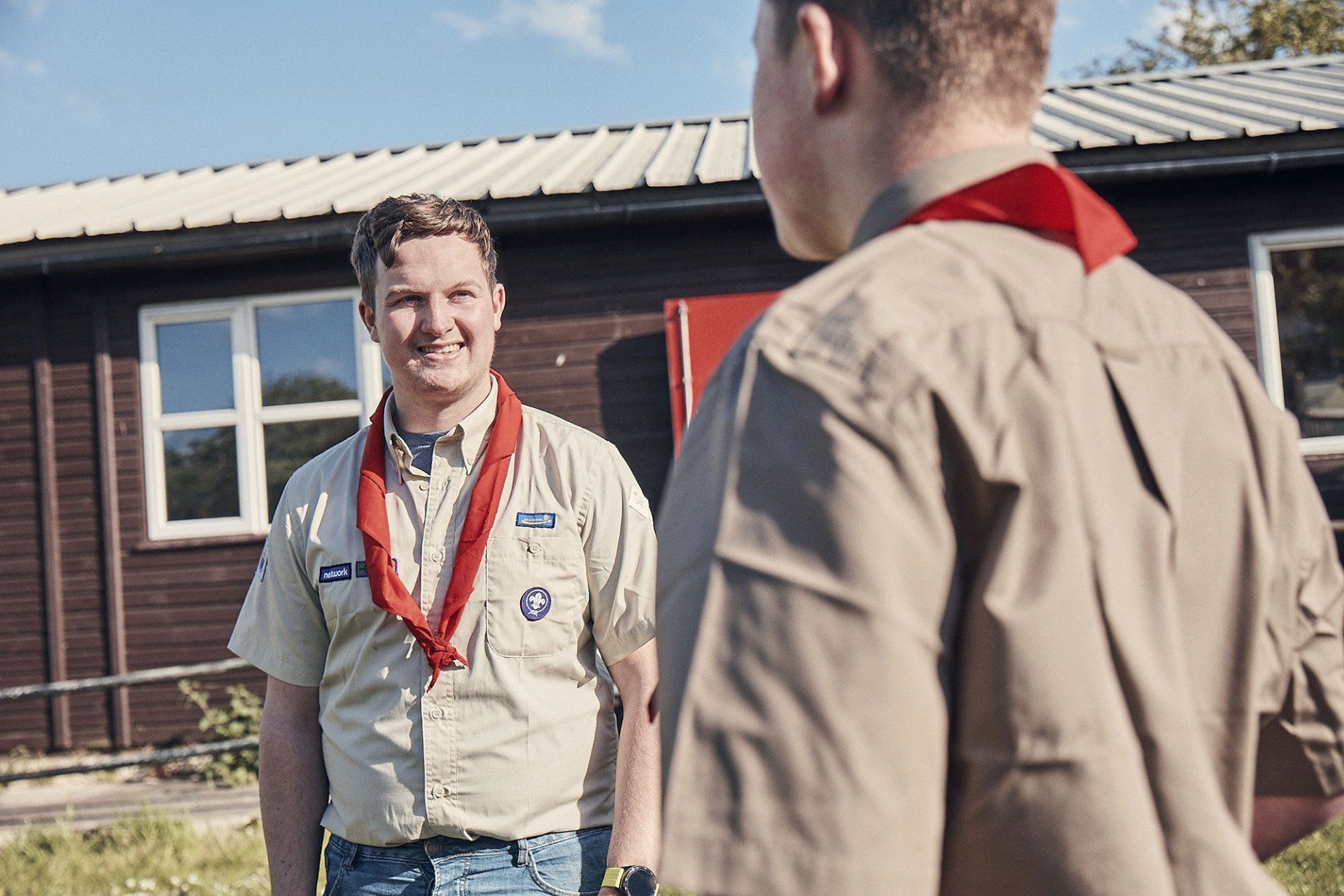 Two boy scouts are standing in front of a wooden building talking to each other.
