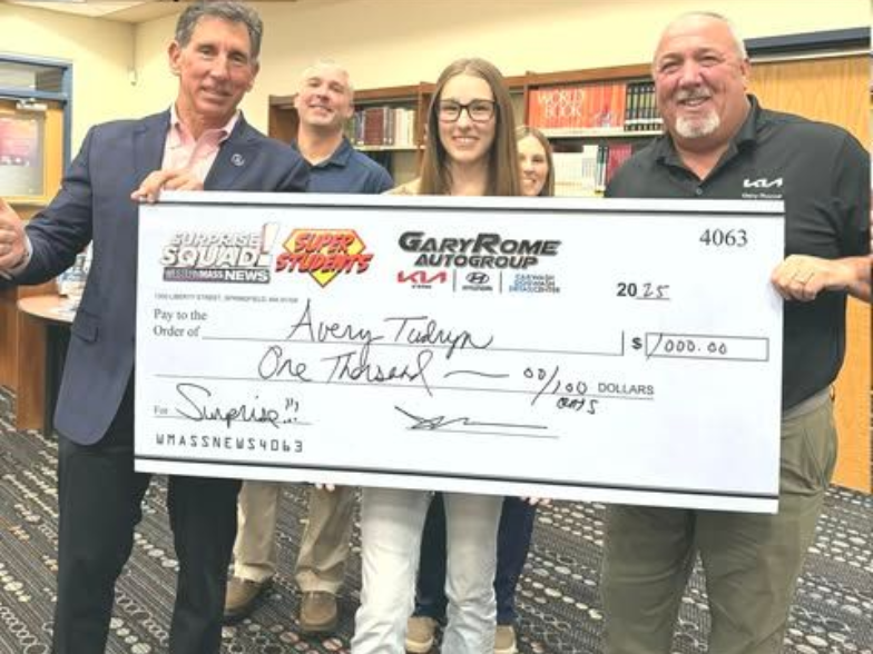 Four people indoors hold an oversized ceremonial check in a library-like room.
