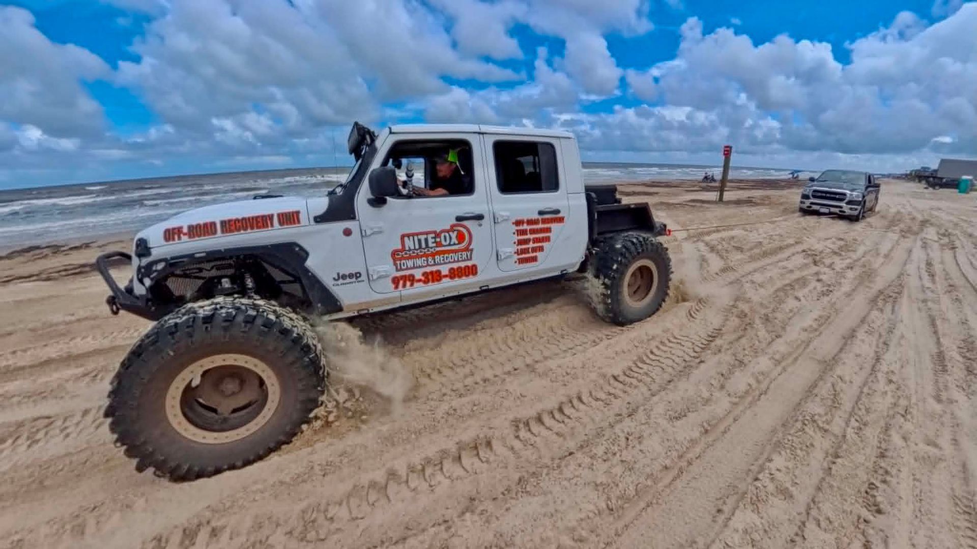 White off-road truck driving on sandy beach under a blue sky, with other vehicles nearby