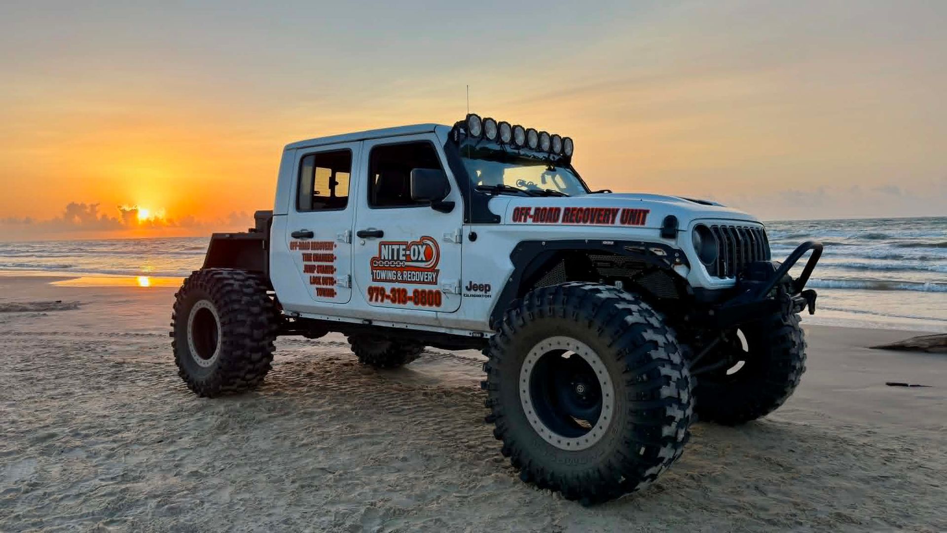 White off-road Jeep on a beach at sunset, with ocean waves and orange sky in the background