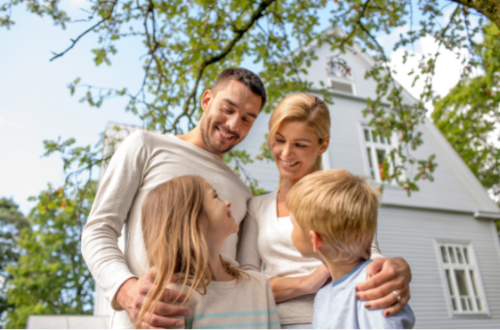 Smiling family embracing outdoors in front of a white house under green tree branches