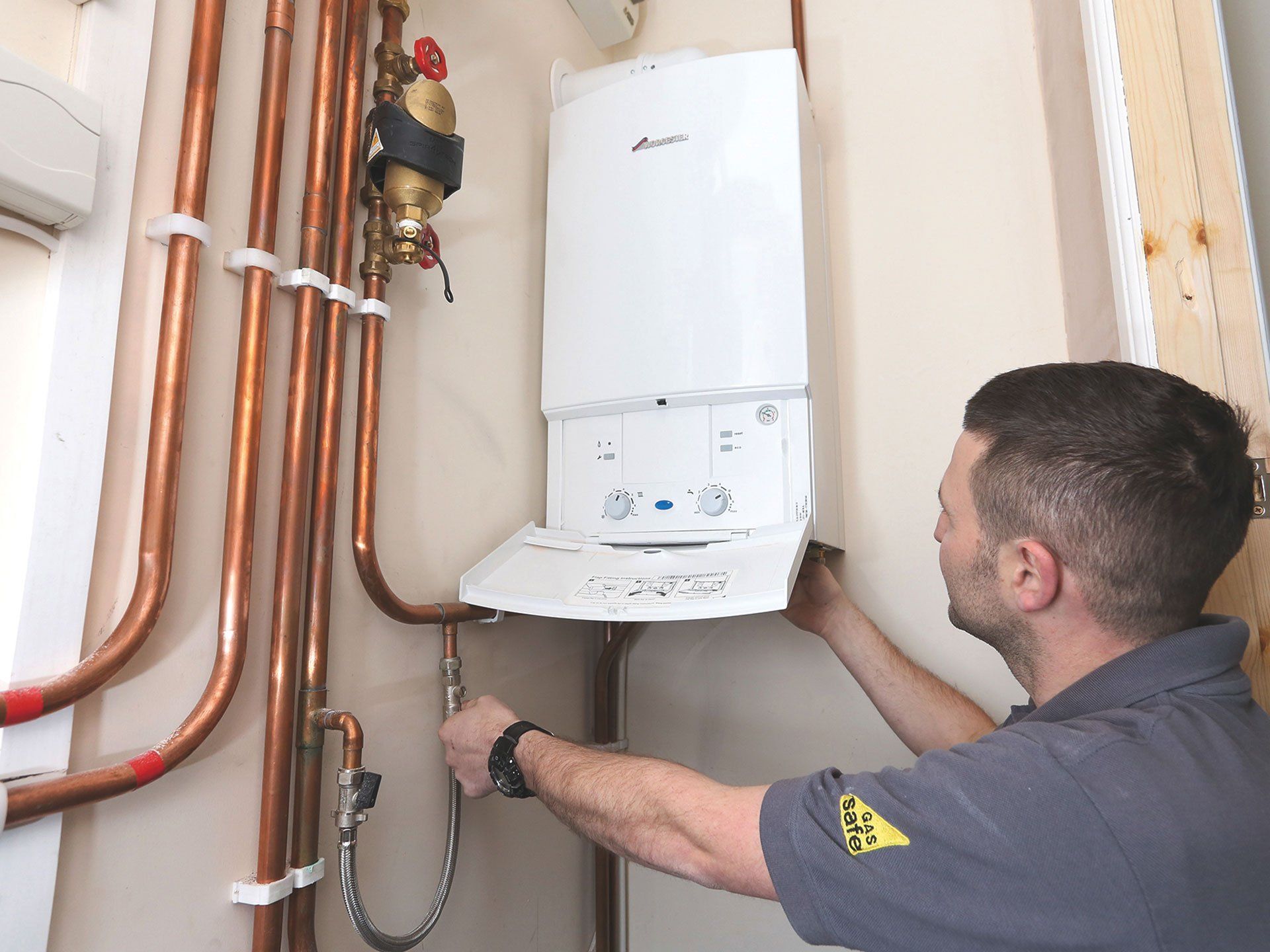 Technician installing a white wall-mounted boiler with copper pipes in a utility room.