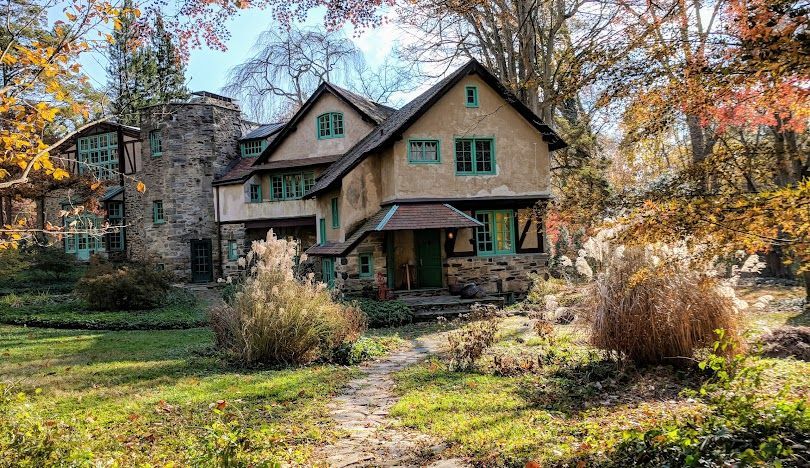 Stone house with green-trimmed windows in an autumn garden, surrounded by colorful trees and shrubs.