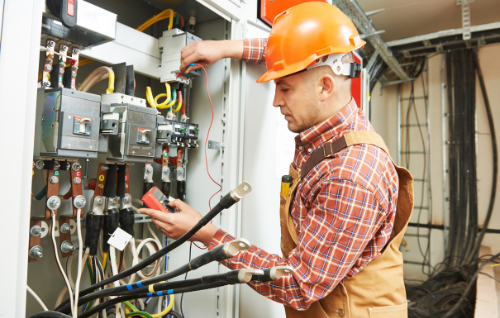 Electrician in orange hard hat inspecting electrical control panel with a multimeter