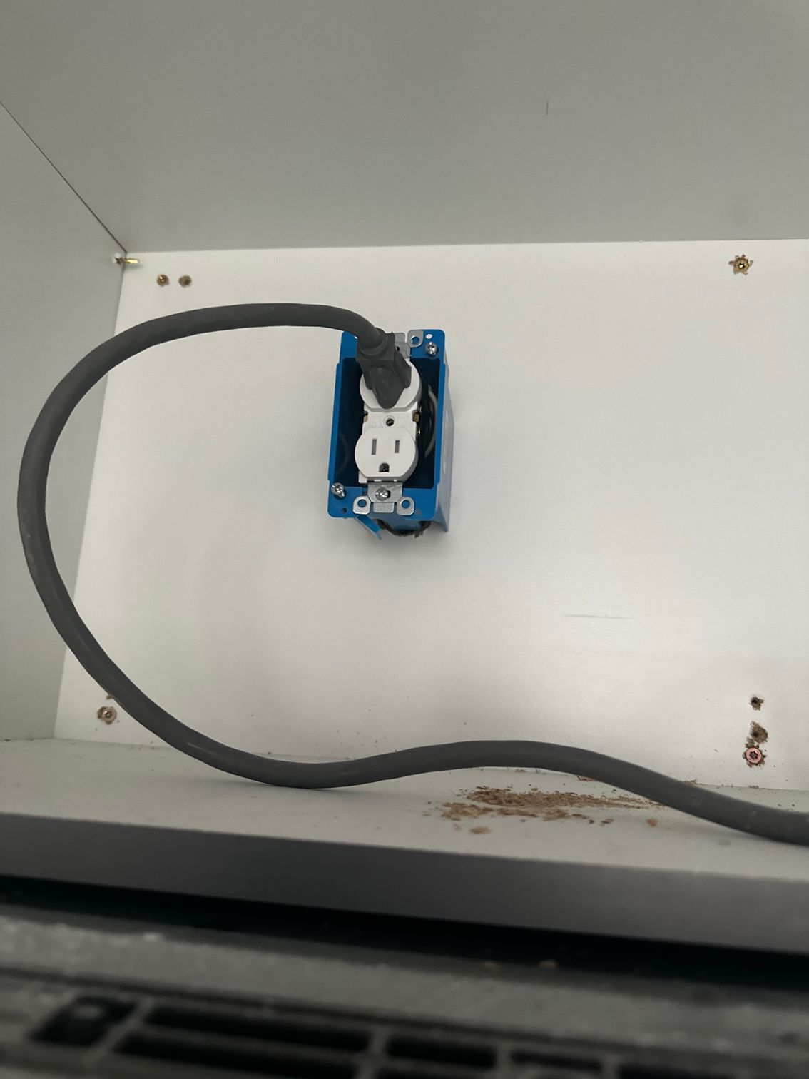 Wall outlet with a black power cord plugged into a blue electrical box on a white panel.