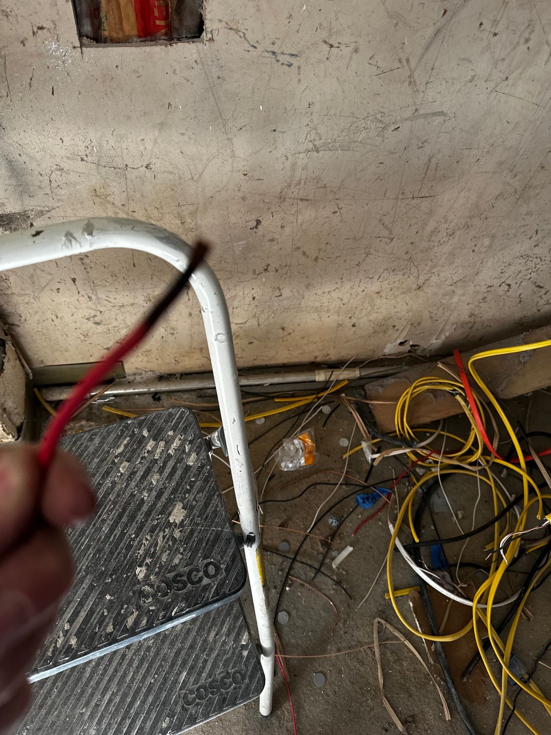 Hand holding red wire near white cable and tangled wires on a dirty floor by a wall