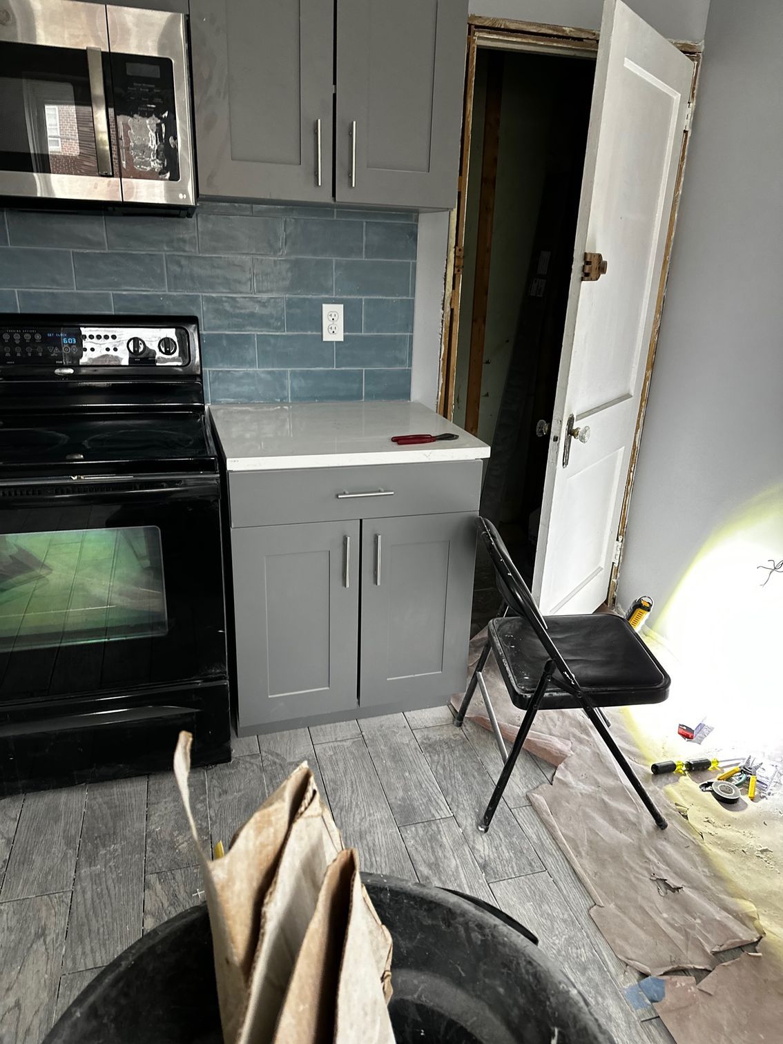 Small kitchen with gray cabinets, black stove, blue tile backsplash, and a chair by an open door.