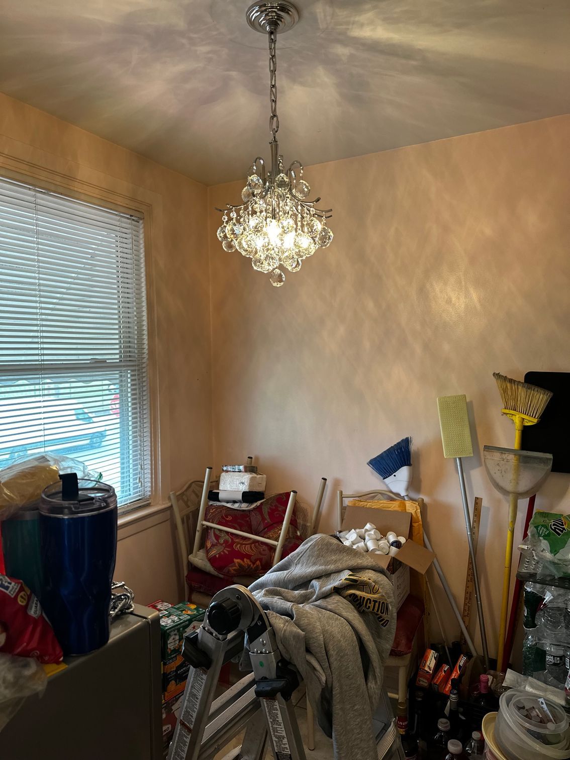 Cluttered room with chandelier, window blinds, and stacked household items against beige walls