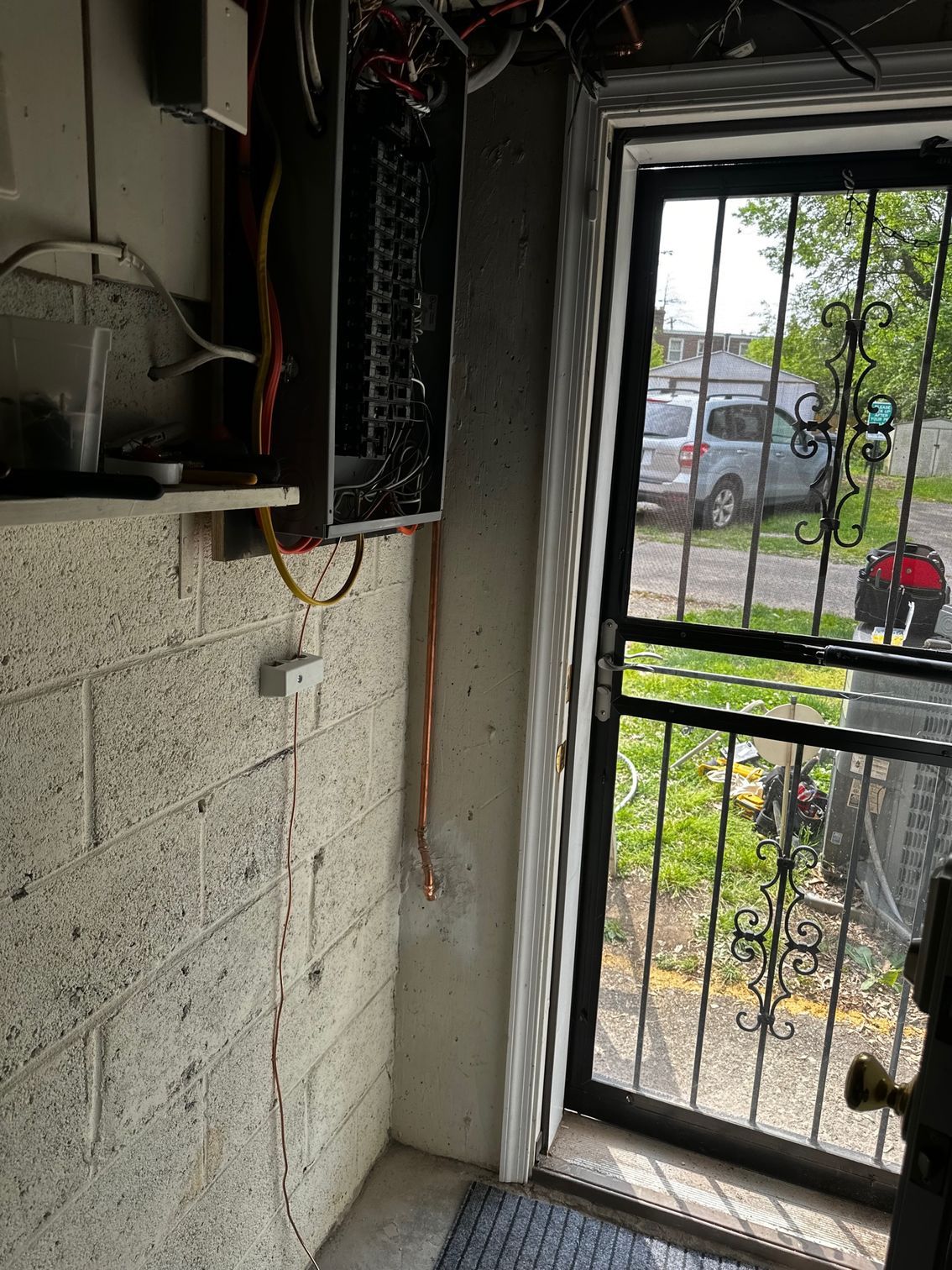 Interior doorway with exposed wiring and a black metal security door looking out to a parked car and greenery.