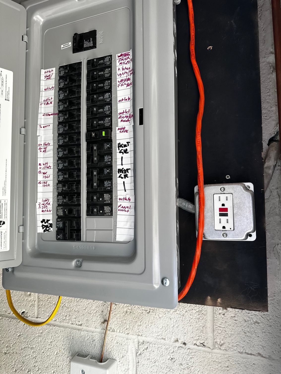 Open electrical breaker panel on a wall with a nearby outlet and orange conduit cable