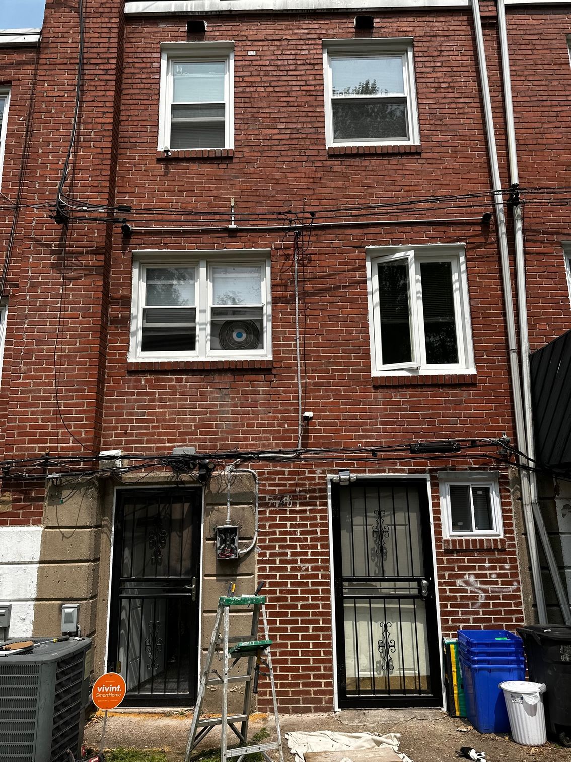 Red brick rowhouse with boarded lower door, broken window, and trash bins in the front alley