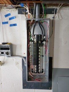Open electrical panel with exposed wiring and circuit breakers in a wall recess