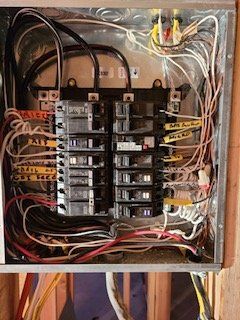 Open electrical breaker panel with tangled wires and labeled circuits inside a wall box