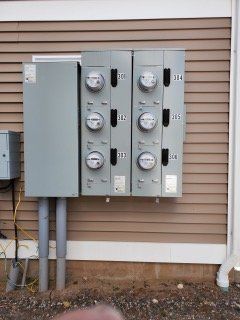 Electric meter panel mounted on a beige house exterior with multiple round meters and conduit pipes.