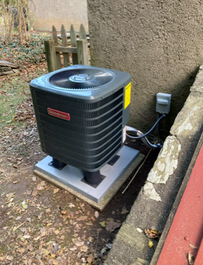 Outdoor air conditioner unit beside a building wall, mounted on a concrete pad with visible wiring.