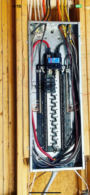 Open electrical breaker panel with wires, circuit breakers, and a central bus bar on a wooden wall.