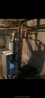 Basement utility room with a water heater, boiler, and exposed pipes against concrete walls.