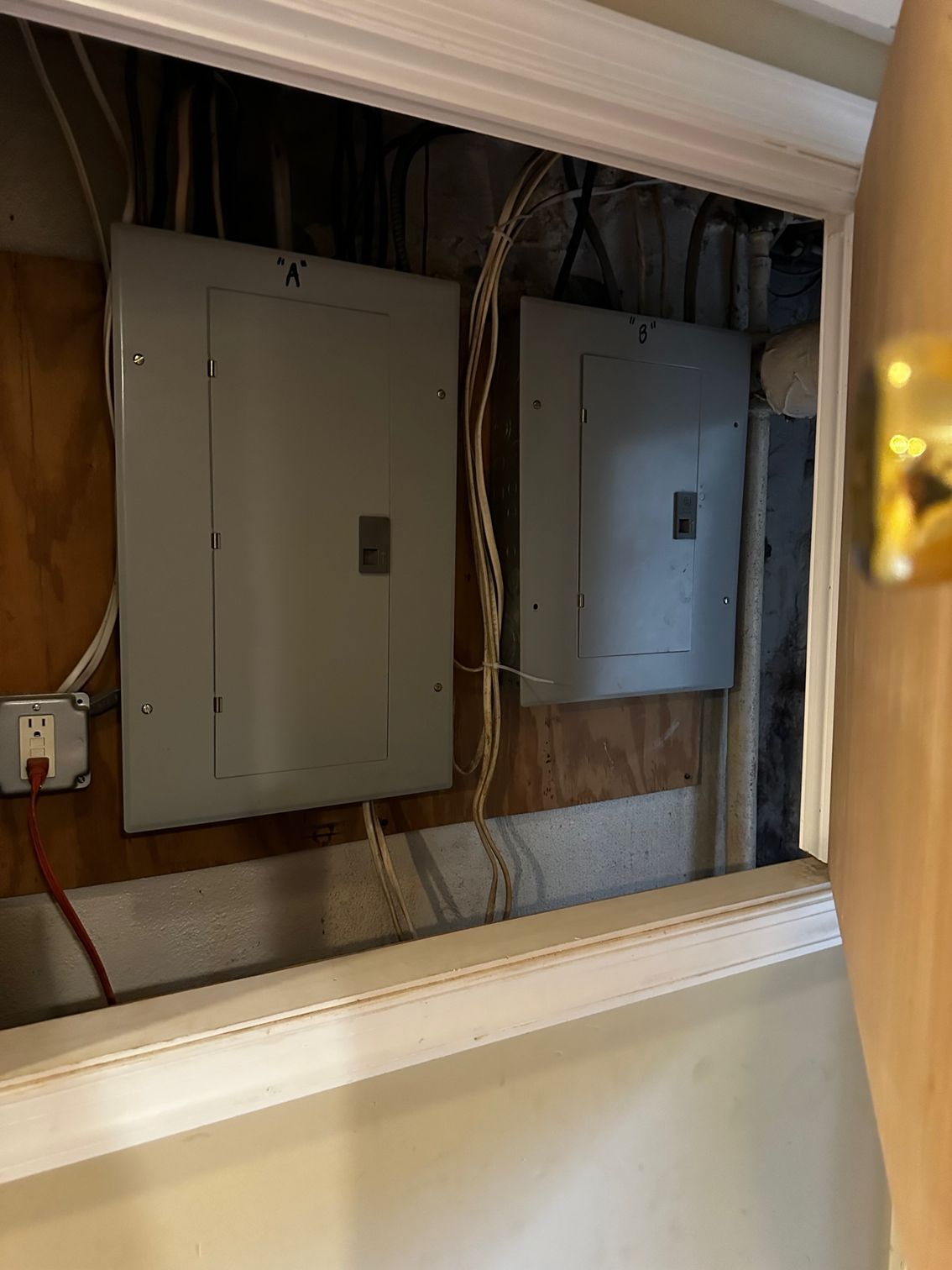 Electrical panels behind an open wall access panel with exposed wires and beige trim.