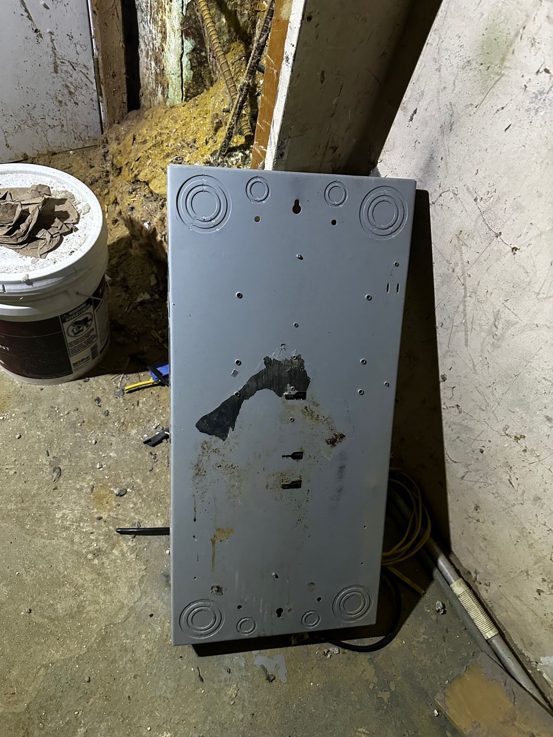 Grey metal electrical panel leaning on a concrete floor in a dirty utility room.