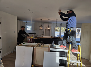 Two workers installing lights in a kitchen under renovation, one on a ladder and one by the island.