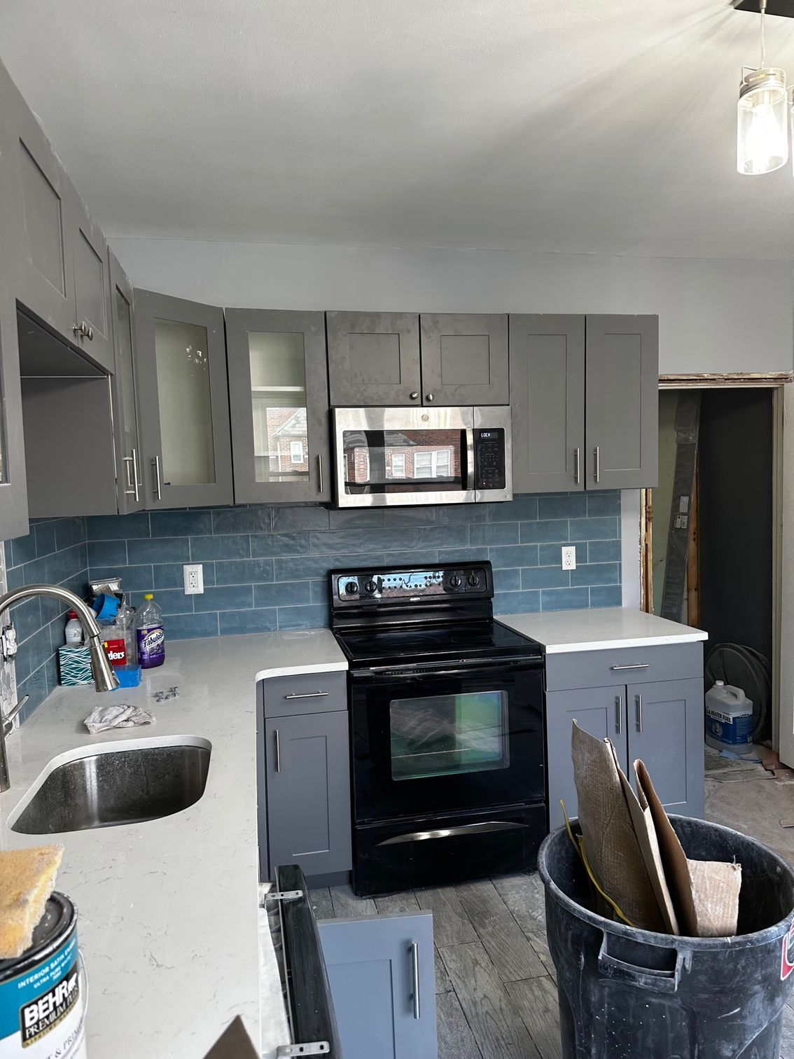 Small kitchen with gray cabinets, blue tile backsplash, black stove, and countertop utensils from a sink-side view