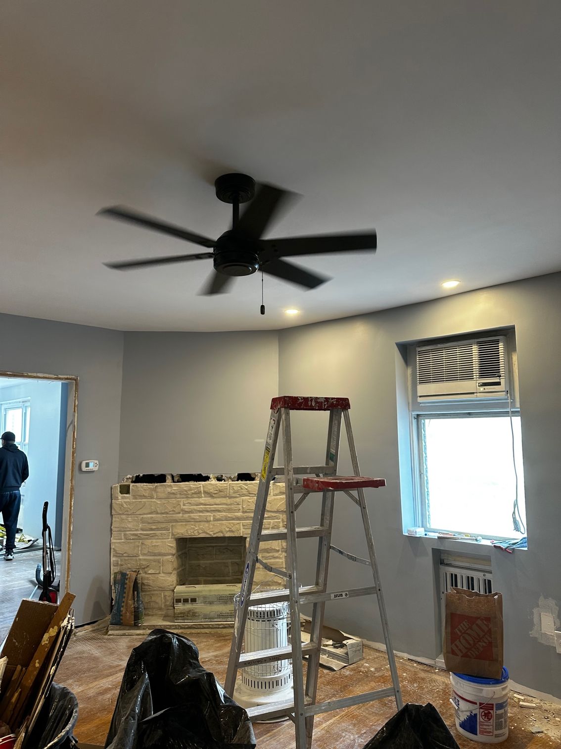 Living room under renovation with ladders, tools, exposed fireplace, and a ceiling fan
