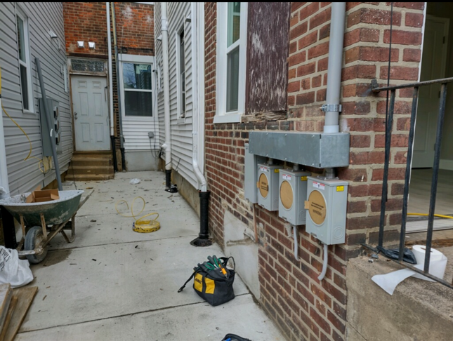 Narrow brick alley beside a house with utility meters, a wheelbarrow, and a black bucket on the concrete path