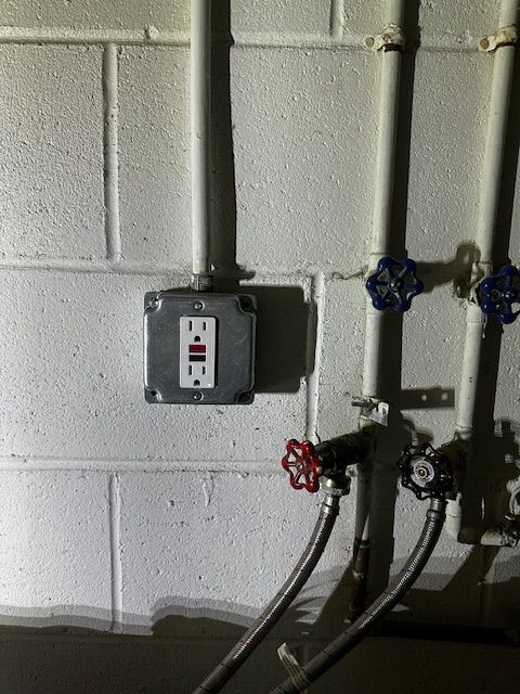 Wall-mounted electrical outlet and shutoff valves on a cinder block wall with exposed pipes.