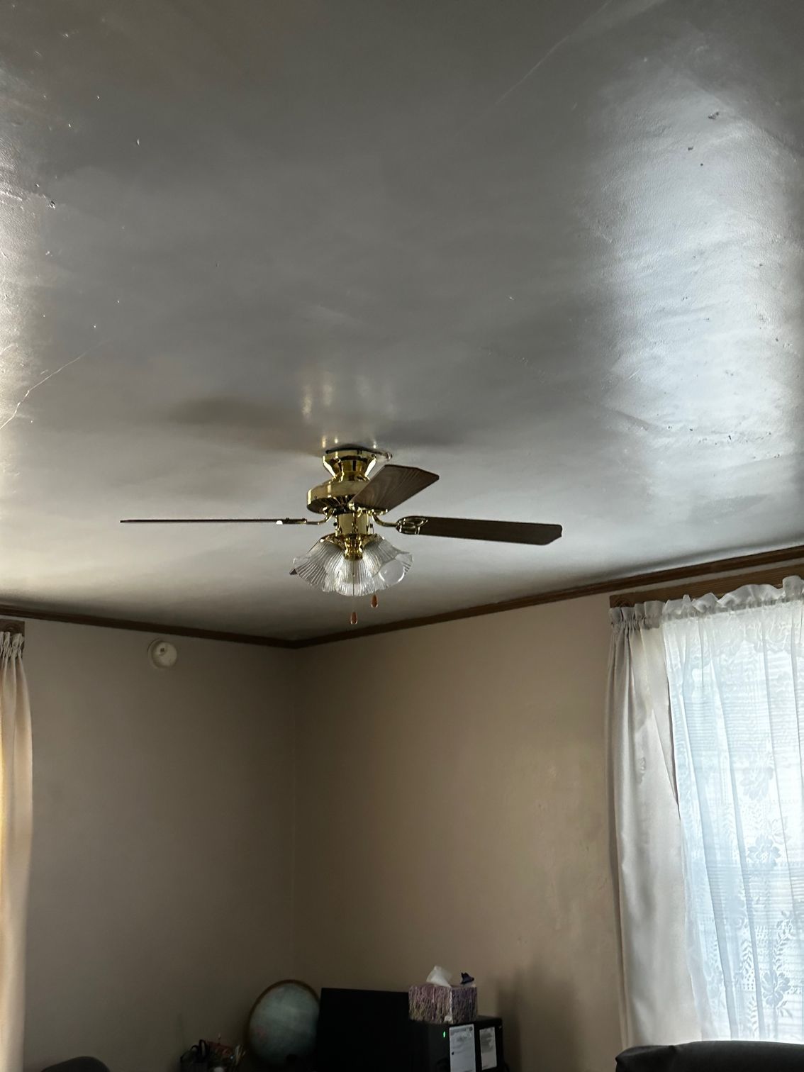 Living room ceiling with a brass fan, beige walls, and a bright window with sheer curtains