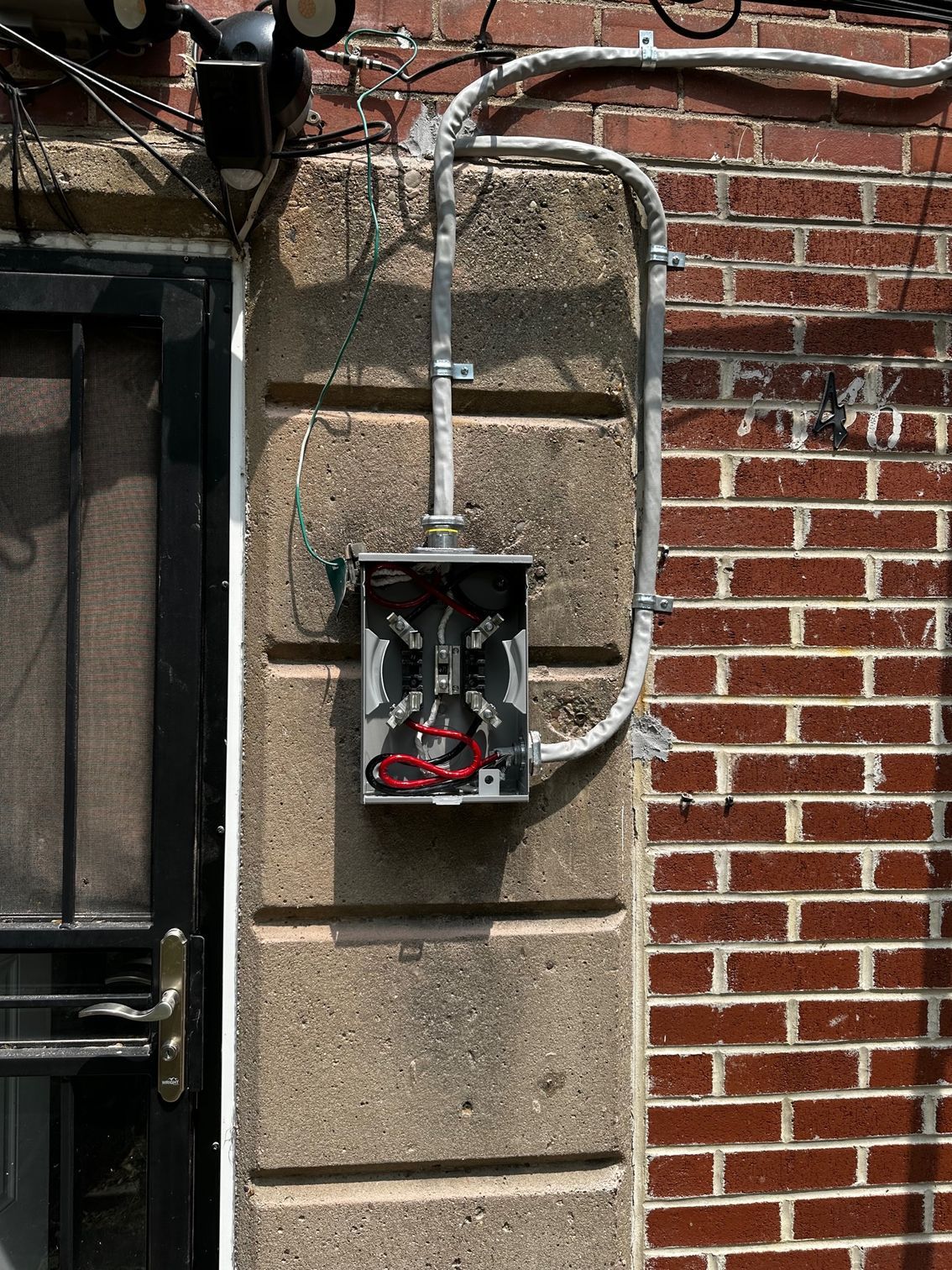Open electrical meter box mounted on a brick wall with exposed wires and conduit.