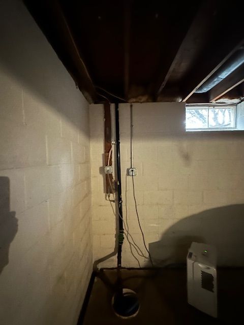 Dim basement utility room with exposed ceiling beams, electrical wires, a window, and a white appliance.