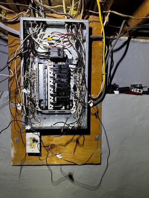 Open electrical breaker panel on plywood with exposed wires and cables