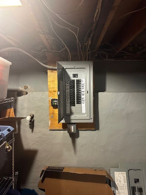 Electrical breaker panel in a basement wall with exposed wires above and storage items nearby
