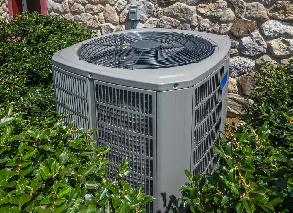 Outdoor air conditioning unit beside a stone wall, partly surrounded by green bushes