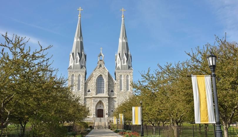 Twin-spired stone church framed by trees under a blue sky, with yellow-and-white banners in the foreground