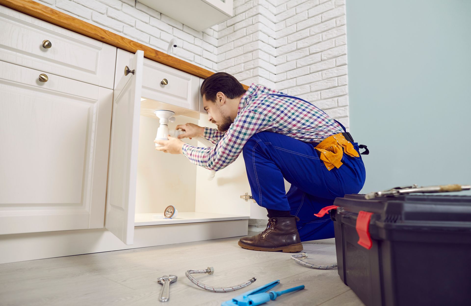 Close-up of plumber, from a plumbing company, repairing the sink with a tool in a bathroom. Person installing under a kitchen cabinet, with tools on the floor and a white tiled backsplash.