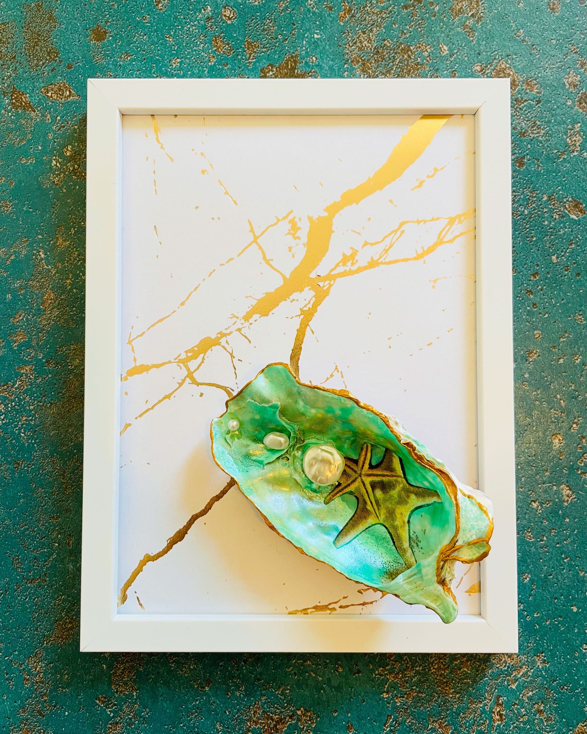 Een foto van een zeegroene schelp met een zeester op wit en gouden achtergrond in een wit kader.