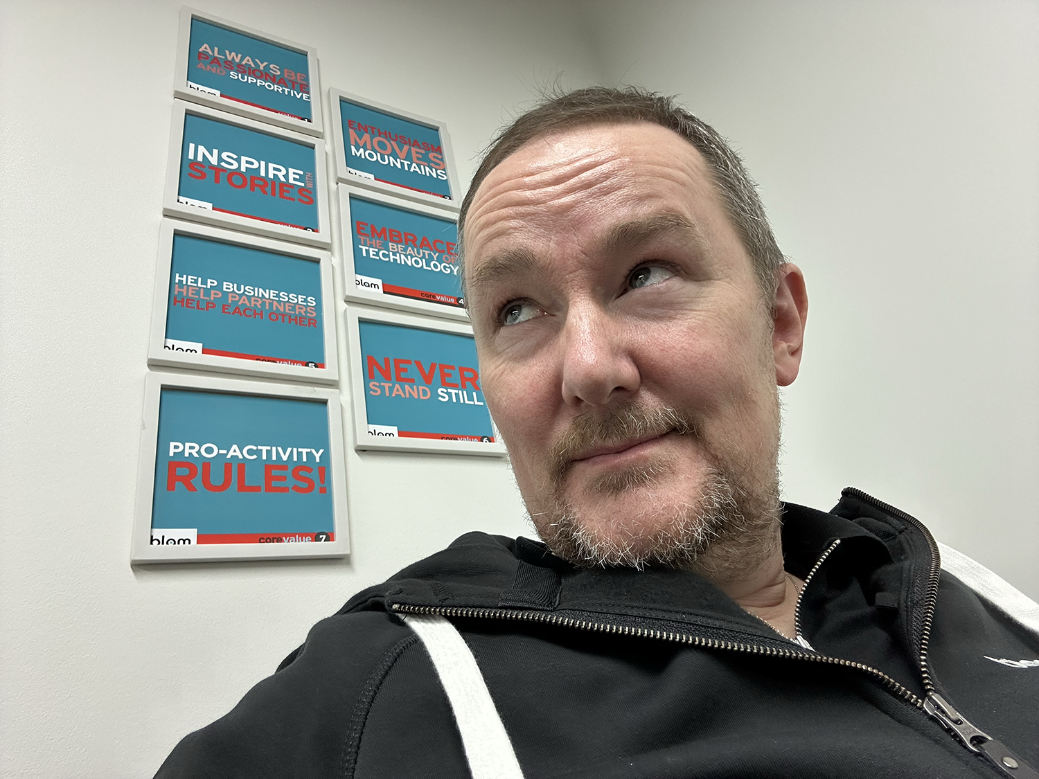 Man in a black hooded jacket looking upward with a thoughtful expression; motivational posters on the wall.
