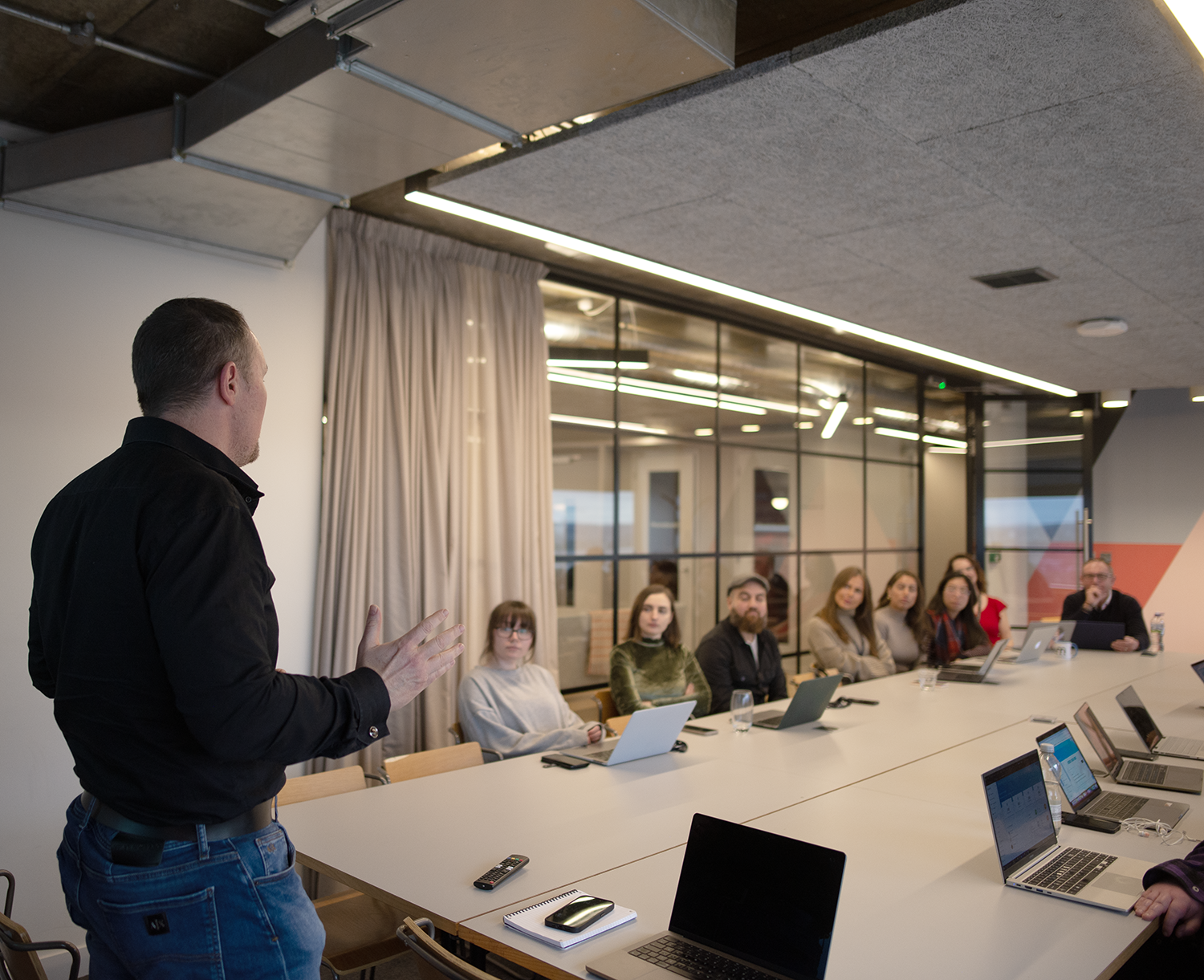 Man presenting to a group seated at a long table in a modern office; laptops, neutral tones.