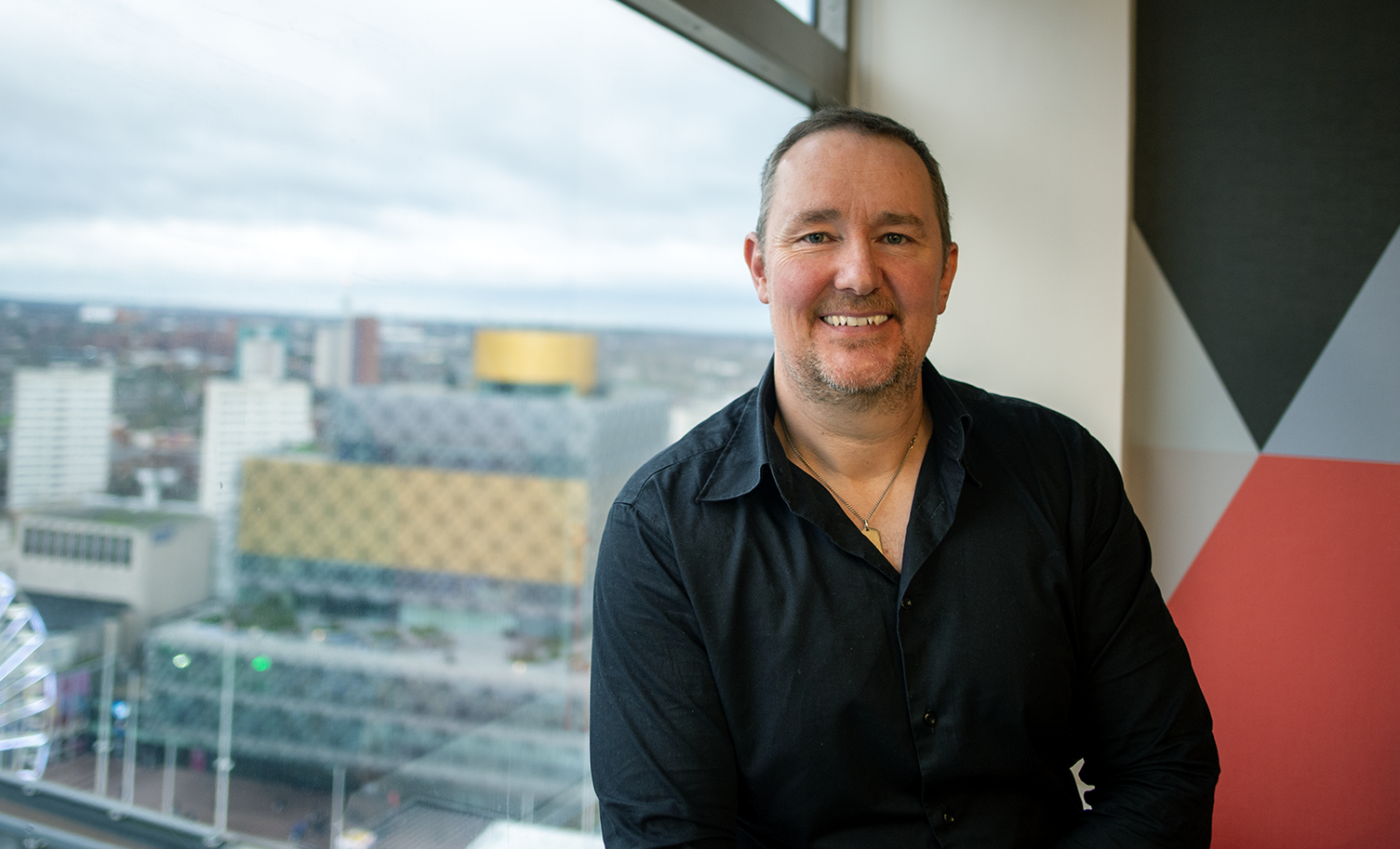Man in black shirt smiles, sits by window with cityscape view. Red and grey geometric wall accent.