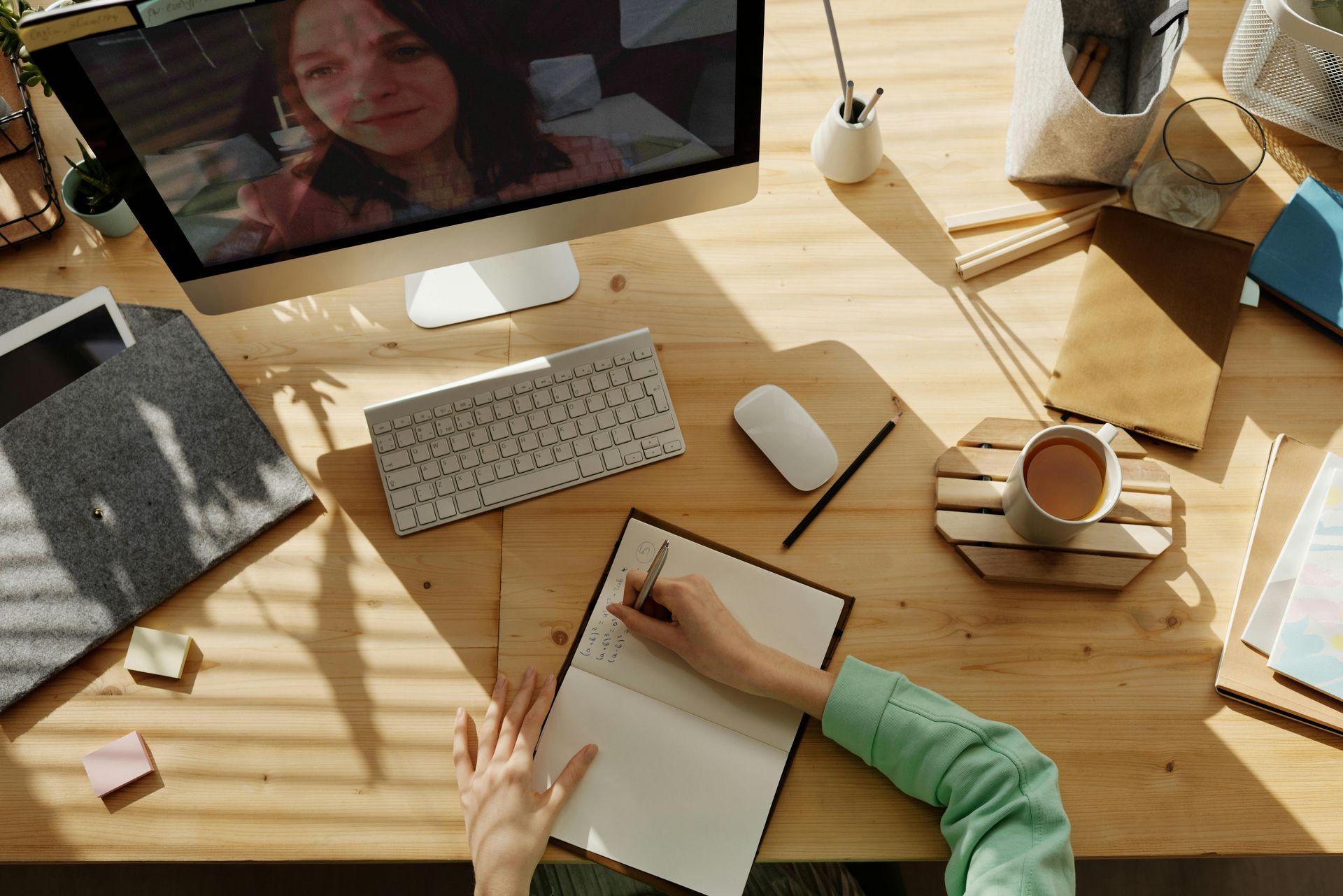 Person at a wooden desk taking notes during a video call with a computer, coffee, and office supplies.
