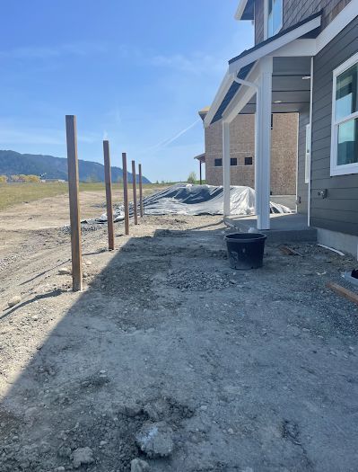 Construction site with fence posts alongside a new house. Gravel ground and blue sky.