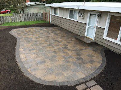 Stone patio with curved border, beside a house with a door and windows, surrounded by dirt and grass.